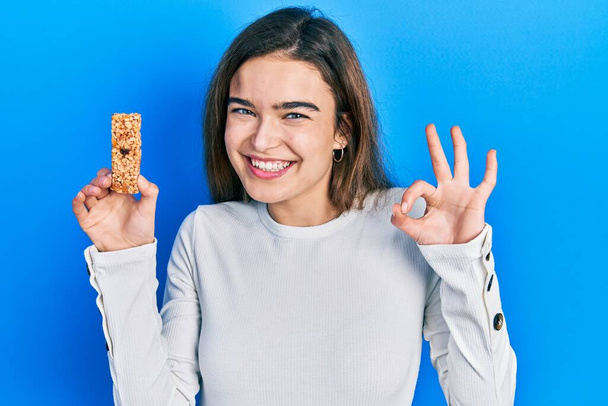 Молодая кавказская девушка ест протеиновый бар в качестве здоровой энергетической закуски делает хорошо знак с пальцами, улыбаясь дружелюбный жест отличный символ  - Фото, изображение