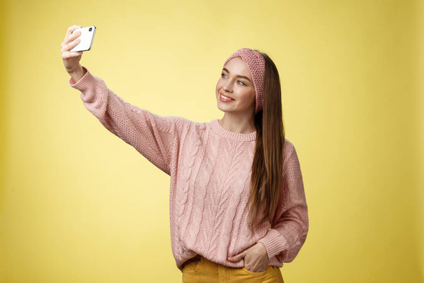 Популярна гламурна молода жінка інтернет-журналістка, яка бере селфі на новий смартфон, розширюючи руку, фотографує себе на жовтому тлі, посміхаючись на екрані мобільного телефону, позуючи нахабною
 - Фото, зображення