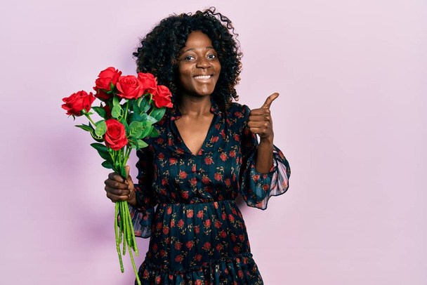 Joven mujer afroamericana sosteniendo ramo de rosas rojas sonriendo feliz y positivo, pulgar hacia arriba haciendo excelente y signo de aprobación  - Foto, imagen