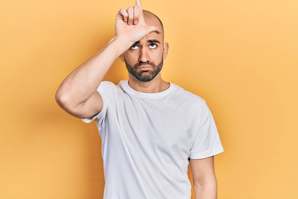 Young bald man wearing casual white t shirt making fun of people with fingers on forehead doing loser gesture mocking and insulting.  - Photo, Image
