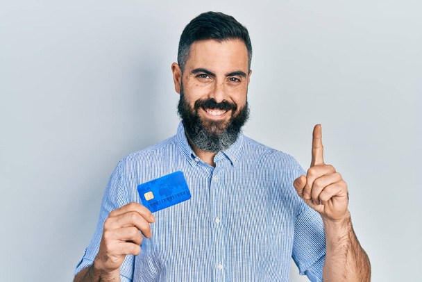 Junge hispanische Mann mit Kreditkarte lächelnd mit einer Idee oder Frage Zeigefinger mit glücklichem Gesicht, Nummer eins  - Foto, Bild