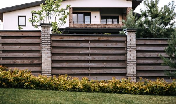 Section of a wooden fence with green plants. Evergreen hedge. Horizontal lines. Brick post. Scandinavian style of backyard. Western border. Home security barrier. Landscaping design. Unedged board. - Photo, Image