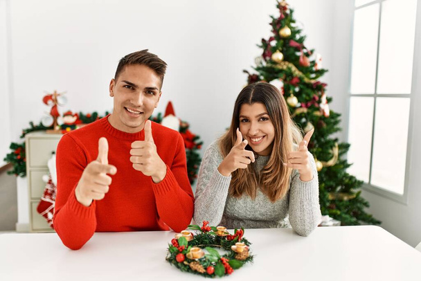 若いヒスパニック系のカップルのクリスマスのカメラに幸せと面白い顔を指を指してテーブルに座っている。良いエネルギーと振動は.  - 写真・画像