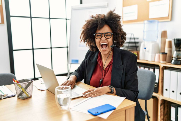 African american woman with afro hair working at the office wearing operator headset angry and mad screaming frustrated and furious, shouting with anger. rage and aggressive concept.  - Fotó, kép