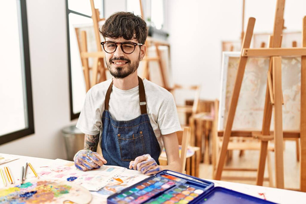 Hispanischer Mann mit Bart im Kunststudio zwinkert in die Kamera mit sexy Gesichtsausdruck, fröhlichem und glücklichem Gesicht.  - Foto, Bild