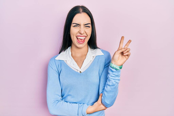 Beautiful woman with blue eyes standing over pink background smiling with happy face winking at the camera doing victory sign. number two.  - Photo, Image