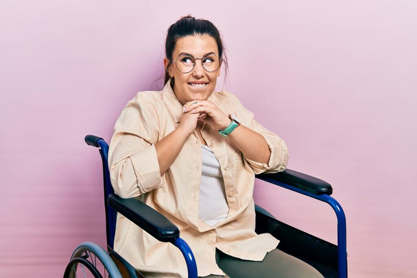 Young hispanic woman sitting on wheelchair laughing nervous and excited with hands on chin looking to the side  - Foto, immagini