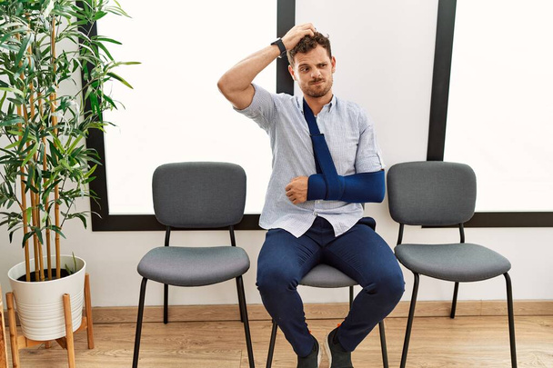 Handsome young man sitting at doctor waiting room with arm injury confuse and wondering about question. uncertain with doubt, thinking with hand on head. pensive concept.  - Foto, imagen