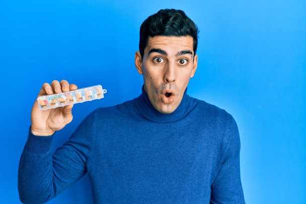 Hombre hispano guapo sosteniendo organizador de píldora asustado y sorprendido con la boca abierta para sorpresa, cara de incredulidad  - Foto, Imagen