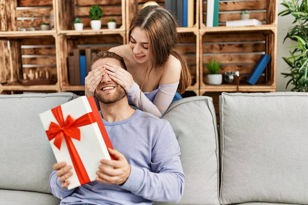 Mujer joven sorprendiendo a su novio con regalo de cumpleaños en casa. - Foto, Imagen