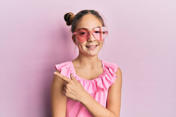 Schönes brünettes kleines Mädchen mit herzförmiger Sonnenbrille lächelt fröhlich und zeigt mit Hand und Finger zur Seite  - Foto, Bild