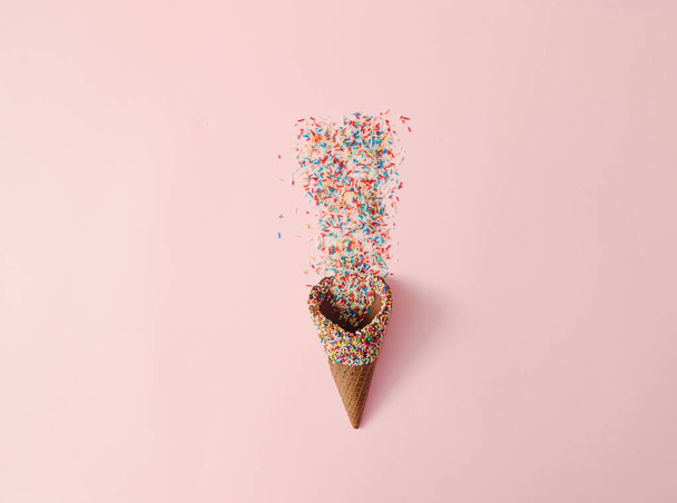 An ice cream cone with colorful crumbs on it and falling out of it on a pastel pink background. Summer sweets creative concept. Colorful ice cream design. - Zdjęcie, obraz