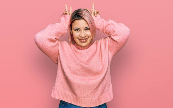 Hispánka s růžovými vlasy na sobě ležérní zimní svetr pózuje legrační a šílený s prsty na hlavě jako zajíček uši, usmívající se veselý  - Fotografie, Obrázek