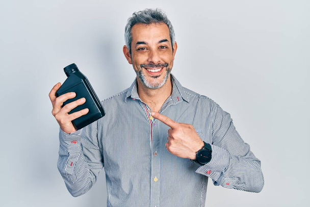 Hombre guapo de mediana edad con pelo gris sosteniendo botella de aceite de motor sonriendo feliz señalando con la mano y el dedo  - Foto, Imagen