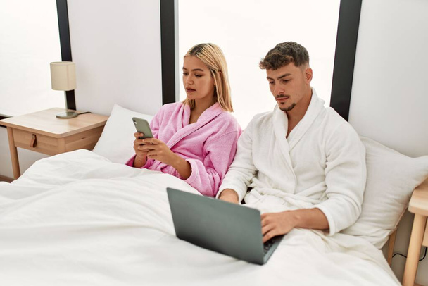 Fiatal kaukázusi pár komoly kifejezés segítségével laptop és okostelefon ül az ágyon otthon. - Fotó, kép