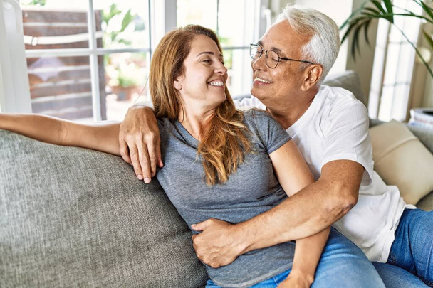 Hispanisches Paar mittleren Alters lächelt glücklich und umarmt zu Hause auf dem Sofa sitzend. - Foto, Bild