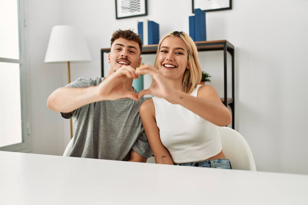 Young beautiful couple wearing casual clothes sitting on the table at home smiling in love showing heart symbol and shape with hands. romantic concept.  - Фото, зображення