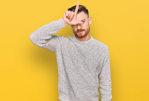 Young redhead man wearing casual winter sweater making fun of people with fingers on forehead doing loser gesture mocking and insulting.  - Foto, immagini