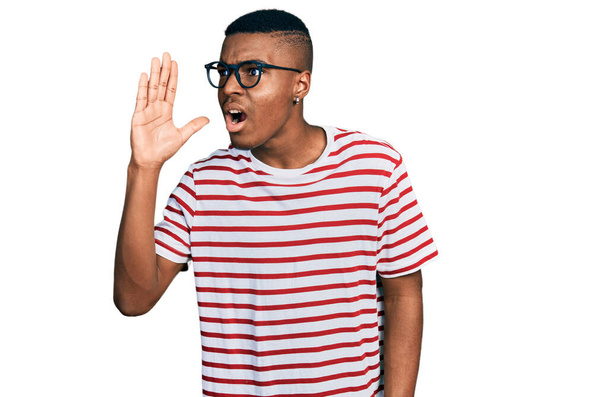 Jeune homme afro-américain portant un t-shirt décontracté et des lunettes criant et criant haut et fort avec la main sur la bouche. concept de communication.  - Photo, image