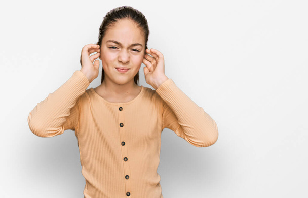 美しいブルネットの女の子は、大きな音楽のノイズのためにいらいらした表情で指で耳を覆うカジュアルなセーターを着ています。聴覚障害の概念.  - 写真・画像