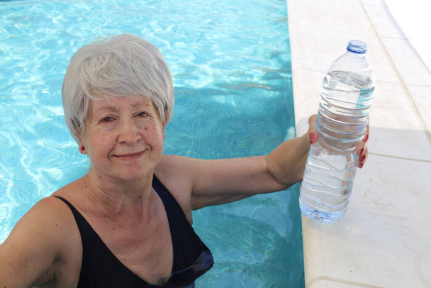 portret van volwassen vrouw in badpak met fles water in het zwembad op zonnige zomerdag - Foto, afbeelding