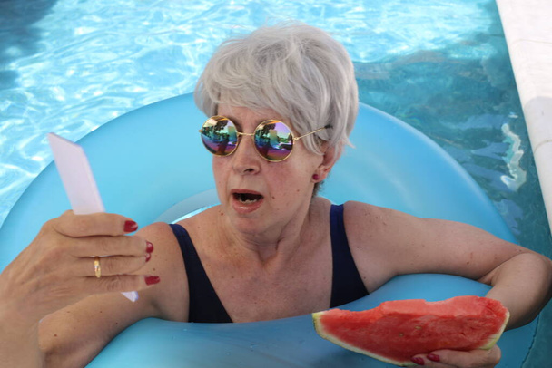 portret van volwassen vrouw in badpak met plak watermeloen met smartphone in zwembad op zonnige zomerdag - Foto, afbeelding