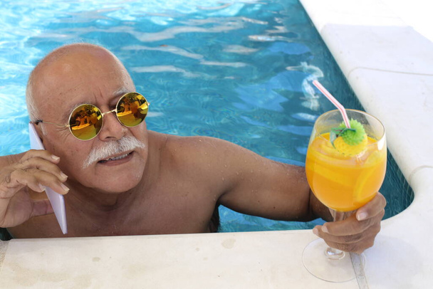 ritratto di uomo maturo con bicchiere di cocktail tropicale che parla per telefono in piscina nella soleggiata giornata estiva - Foto, immagini