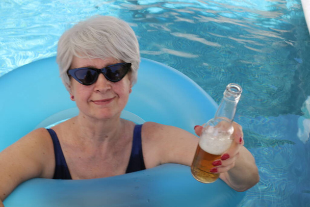 портрет зрелой женщины в купальнике с надувной трубкой, пьющей пиво в бассейне в солнечный летний день - Фото, изображение