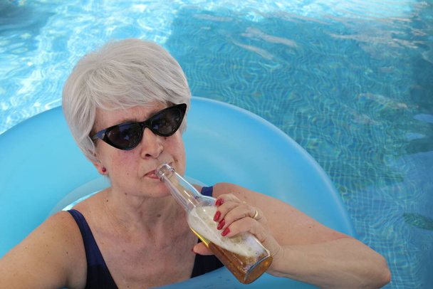 日当たりの良い夏の日にプールでチューブ飲料ビールを飲みながら水着で成熟した女性の肖像画 - 写真・画像