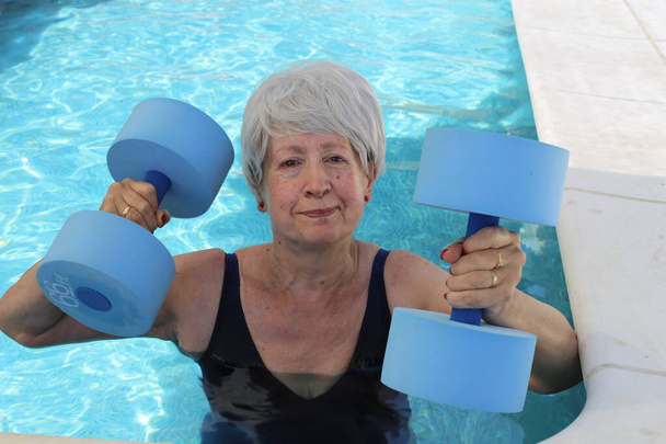 portrait of mature woman in swimsuit with barbells in pool on sunny summer day - Photo, Image