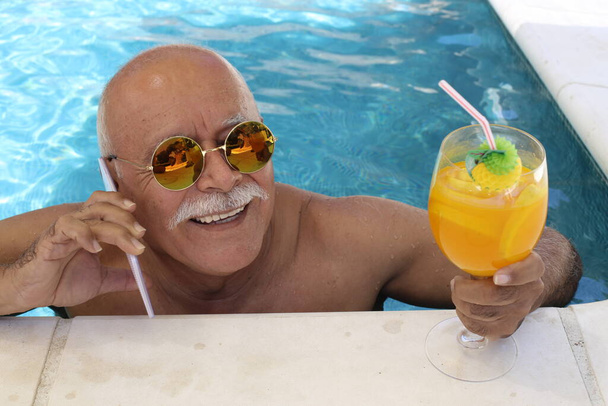 Porträt eines reifen Mannes mit einem Glas tropischem Cocktail, der an einem sonnigen Sommertag im Pool telefoniert - Foto, Bild