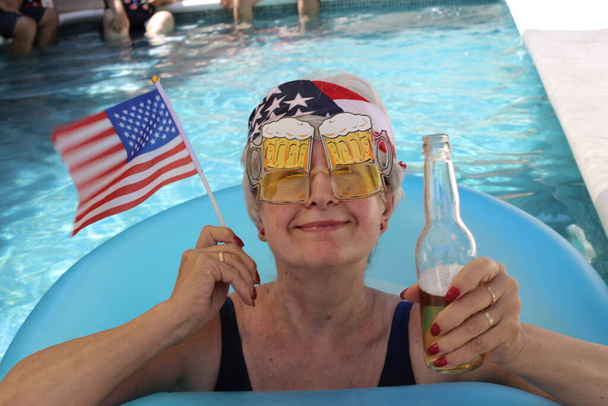 ritratto di donna matura in costume da bagno con bandiera USA, banda e birra in piscina nella soleggiata giornata estiva - Foto, immagini