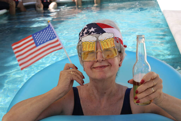 portré érett nő fürdőruhában USA zászló, zenekar és sör medencében a napos nyári napon - Fotó, kép