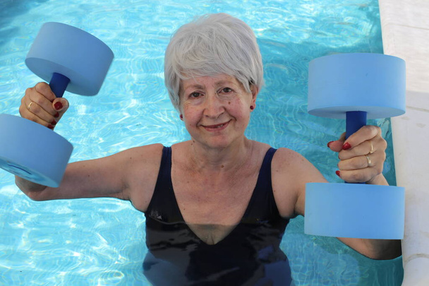 portrait of mature woman in swimsuit with barbells in pool on sunny summer day - Photo, Image