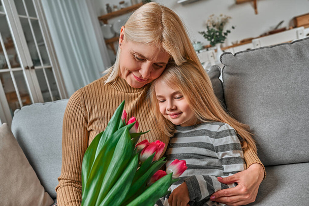 Grandma, this gift is for you! Little beautiful blonde granddaughter gives her grandmother a bouquet of pink tulips. The concept of family, respect, generation, mother, upbringing. - Valokuva, kuva
