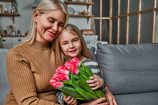 I love you grandma! Grandmother and granddaughter sitting embracing on the couch holding pink tulips. Spring, celebration, mother and woman's day concept - 写真・画像