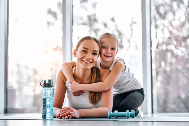 Happy little girl hugging her beautiful sporty mother while lying mats on training. Healthy lifestyle concept - Fotoğraf, Görsel