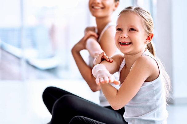 Portrait focused on little daughter and blurred sporty mother in sportswear, making shoulder and hand warm up before training in gym. While exercising woman and girl looking at camera and smiling. Wellbeing concept - Фото, зображення