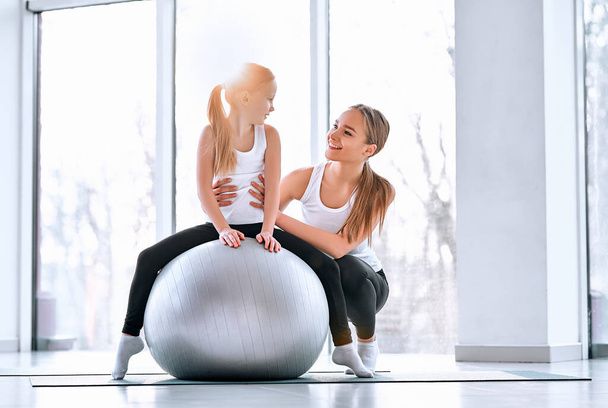 Full lenght shot of happy family on training in gym. Mom helps daughter doing exercises on fit ball. Welness concept - Foto, Imagem