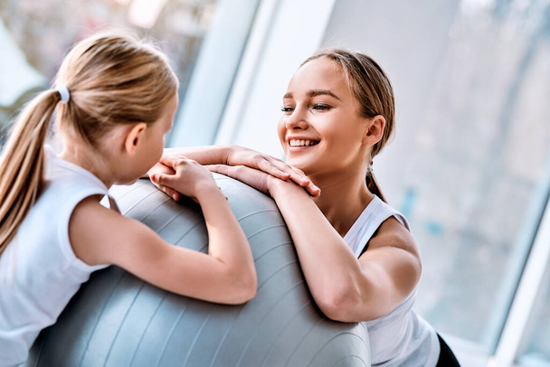 Beautiful young mother sits together with her daughter on the floor near fitball at the gym. Healthy lifestyle concept - Foto, Imagem