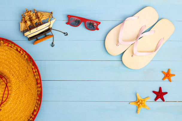 Zomertijd op een koel uitzicht op beschilderde lichtblauwe houten tafel met zonnebril, slippers, zeilboot, hoed en zeester - Foto, afbeelding