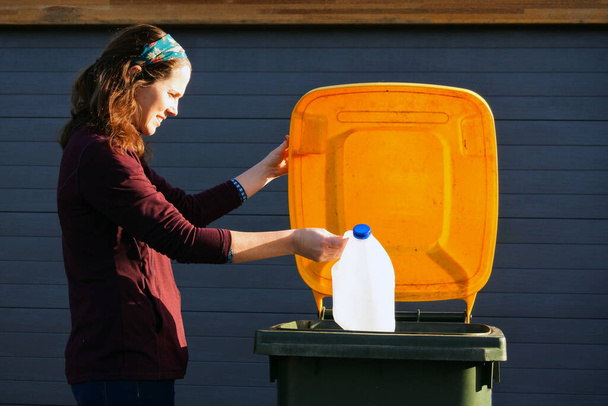 Yetişkin bir Avustralyalı kadın (30-40 yaş) atık toplama gününde evinin dışındaki sarı geri dönüşüm kutusuna plastik bir süt şişesi koyar. - Fotoğraf, Görsel