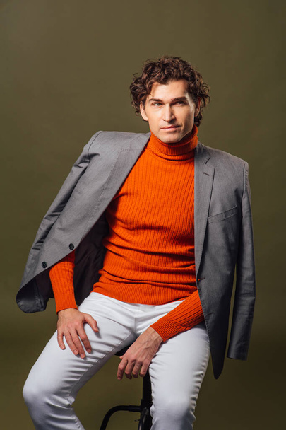 Portrait of a tall handsome man dressed in orange turtleneck, grey jacket and white jeans posing on the green background - Fotoğraf, Görsel