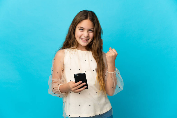 Μικρό κορίτσι πάνω από απομονωμένο μπλε φόντο με το τηλέφωνο στη θέση νίκη - Φωτογραφία, εικόνα