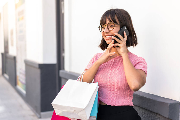 Молодая брюнетка в городе держит сумки с покупками и звонит другу на мобильный телефон - Фото, изображение