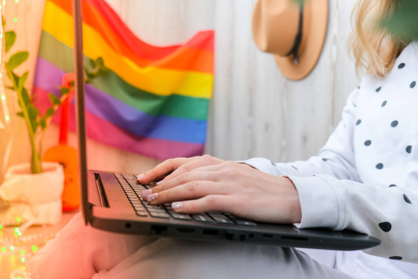 Young millennial hippie woman sitting on balcony using laptop. LGBTQ rainbow flag on background. Online education tuition. Work from home. Modern office. Blonde hair. Stay home - 写真・画像