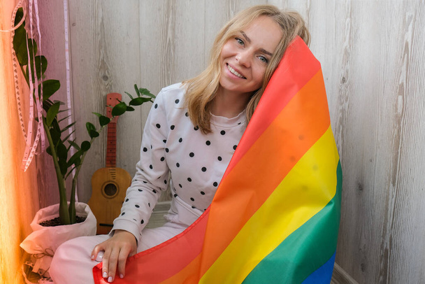 Young blonde millennial hippie woman with rainbow LGBTQ flag at home balcony. Peace and freedom the symbol of LGBT, spending and sharing loving time. Support LGBTQ community. Young lesbian woman - Foto, afbeelding