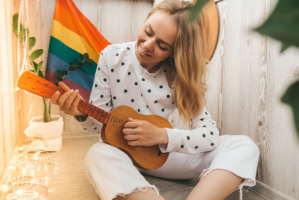 Young millennial hippie woman sitting on balcony play guitar. Music lesson and singer sitting. LGBTQ rainbow flag on background. Online music tuition. Playing instrument and enjoying songs and leisure activity indoor. Blonde hair. Stay home entertain - Фото, зображення