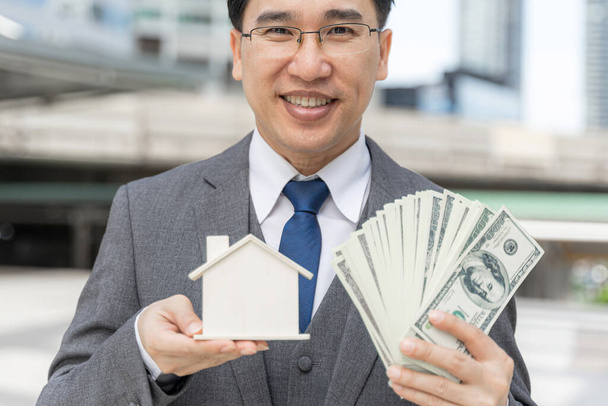 Retrato asiático hombre de negocios con billetes de dólar y la casa modelo en el distrito de negocios, teléfono inteligente en la mano - estilo de vida concepto de la gente de negocios - Foto, Imagen