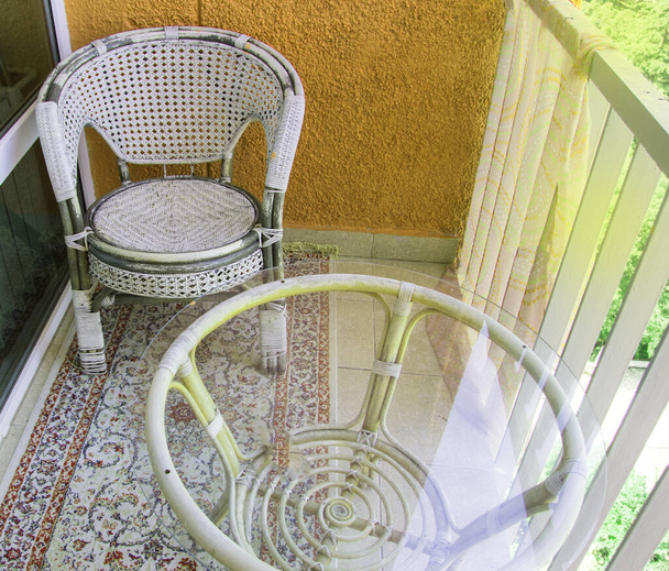 Komfortní posezení na balkóně s ratanovým proutěným křeslem, průhledným skleněným stolem a kobercem, domácí rekreační koncept. - Fotografie, Obrázek